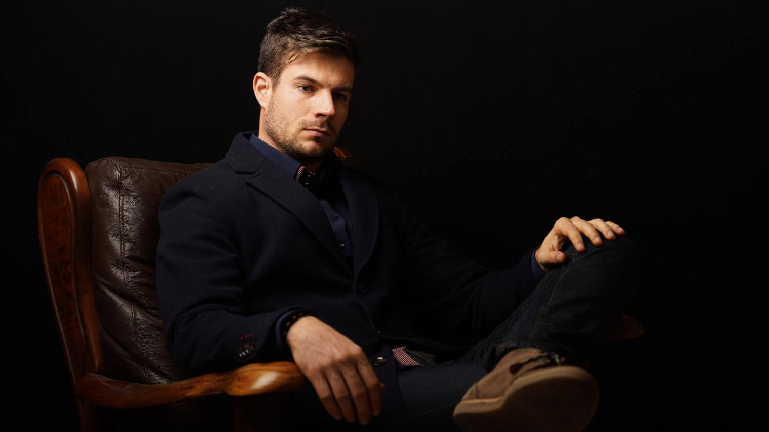 Man sitting in a leather armchair wearing a dark blazer and shirt against a black background, looking relaxed and contemplative.
